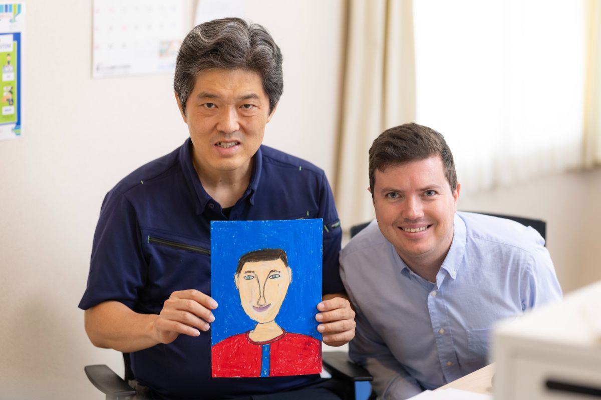 ポップな色使いがとてもかわいい嶋田さんのイラスト。手に持っているのは写真右の社員の方を描いたものです。絵を描くのにかかる時間を聞くと、「1日もかからない（笑）」と笑顔で教えてくれました（写真左が嶋田さん）